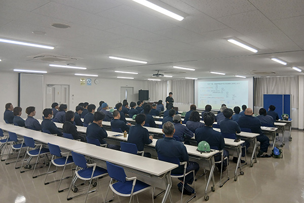 広い部屋に大人数が整列・着席し話を聞いている様子